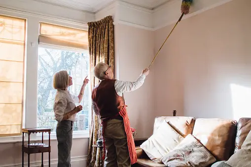 Older man using an extendable duster to dust the ceiling with older woman standing beside him
