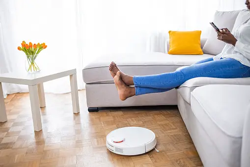 Woman sitting on couch with raised legs with automated vacuum near the couch