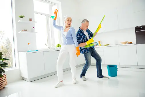 Older man and woman posing in an upright position with cleaning supplies in a kitchen. Man holding a mop like a guitar and woman holding a spray bottle above her head