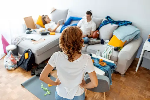 Rear view of mom with hands on hips looking at teenaged son and daughter lounging on couch in a messy living room 