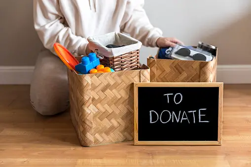 Hands holding two baskets of donated items with chalkboard placed in front, stating 'to donate'