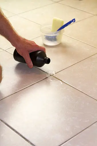 hand pouring bottle of hydrogen peroxide onto tiled floor to clean grout